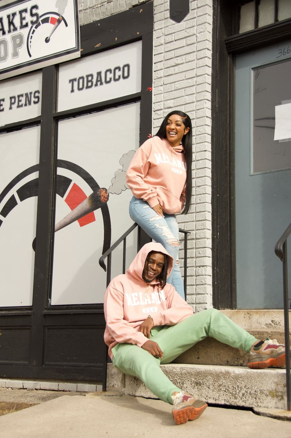 Two models in blush Melanin CLO hoodies on storefront steps