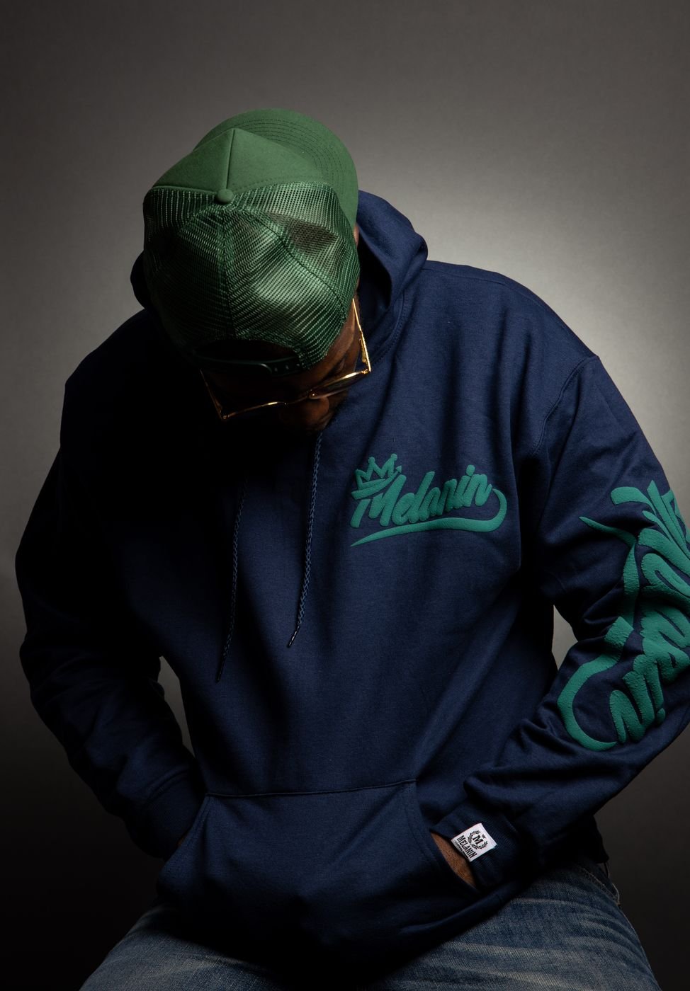 Model in a navy Melanin script hoodie and green cap looking down, studio lighting