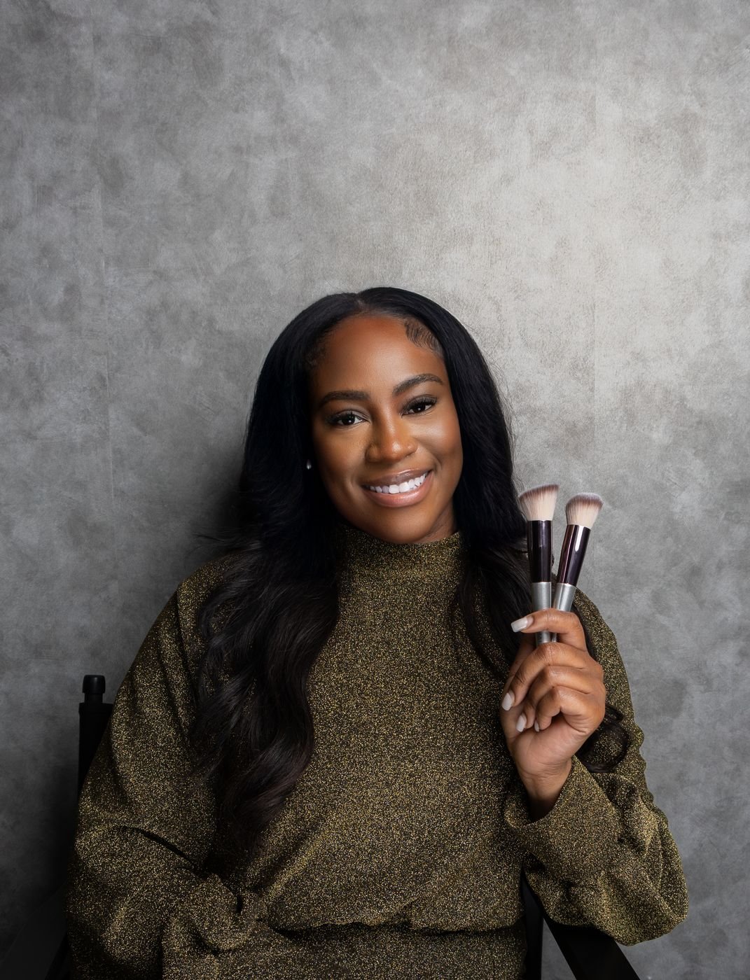 Seated personal-brand headshot holding makeup brushes on a grey backdrop